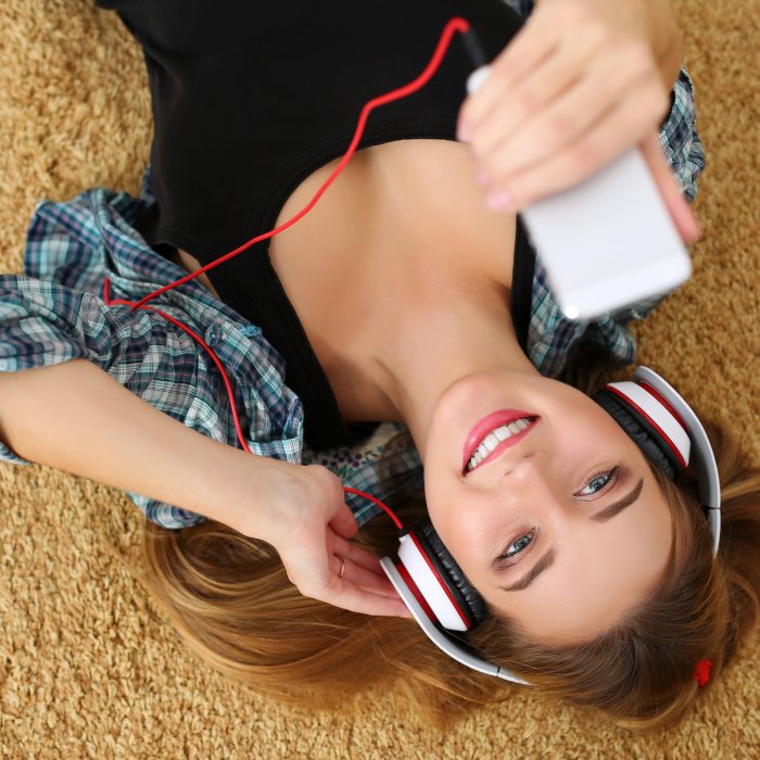 Beautiful blonde smiling woman lying on carpet floor wearing headphones, holding cell phone and listening music portrait. Modern urban life, audio book, radio or education concept